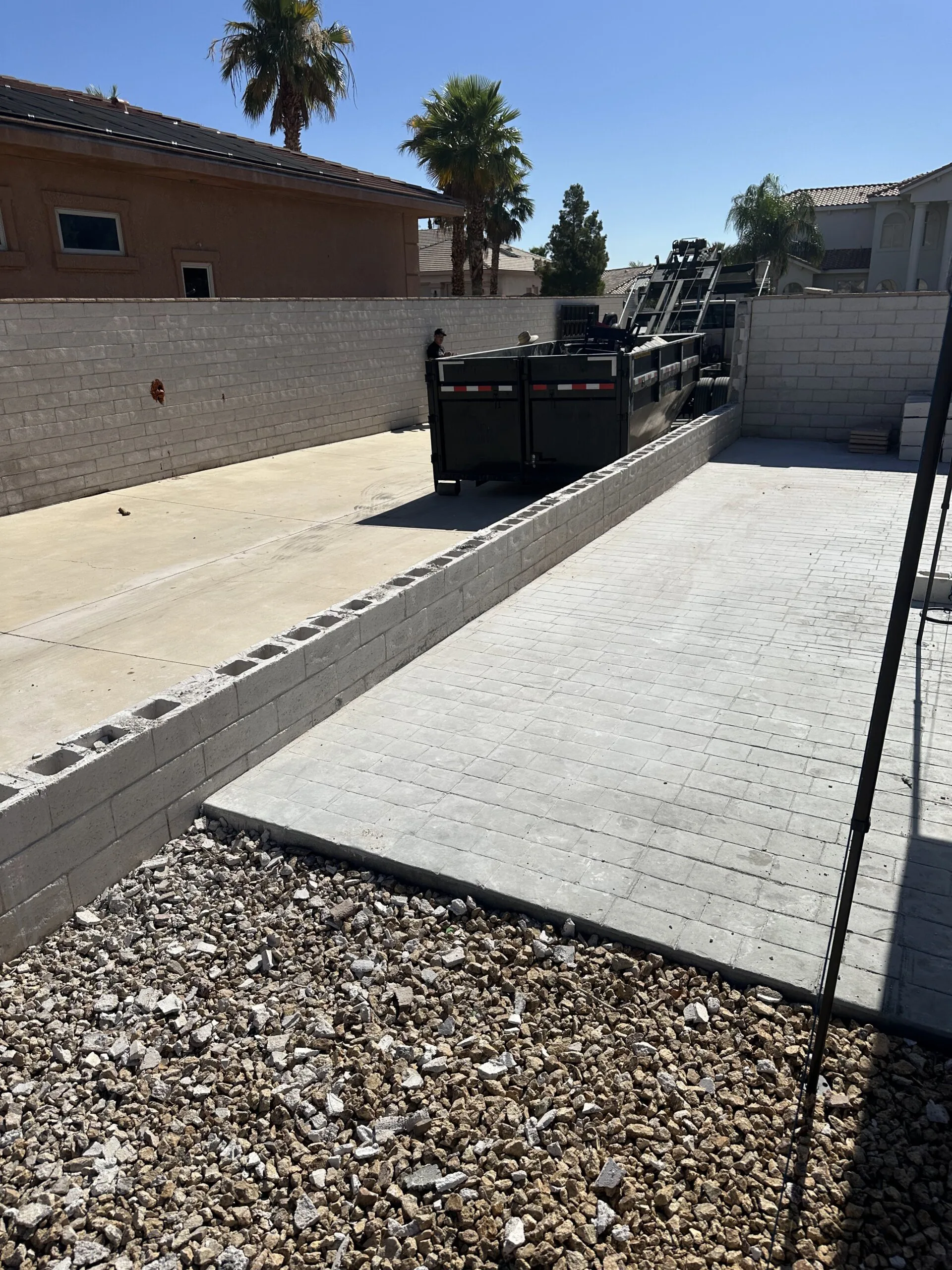 Black dumpster being placed in a walled residential driveway.