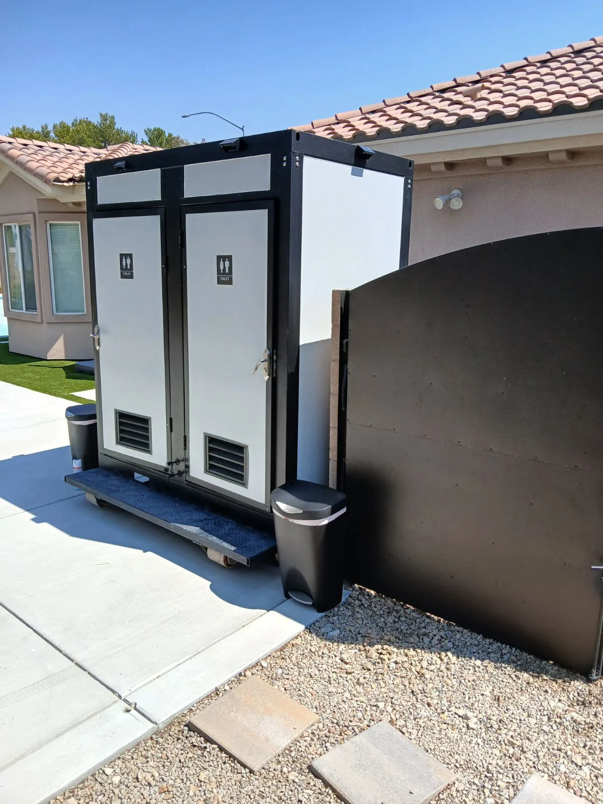 "Dual portable restroom trailer set up in a residential driveway, with trash bins placed outside."